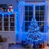 A decorated Christmas tree with blue lights and presents underneath, in a room with a bookshelf and curtains. Produktbild-Vorschau