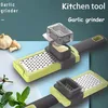 A close-up marketing image of a garlic and ginger grater with a collection container, set against a teal and light blue background. A whole garlic bulb and some herbs are visible in the foreground. Produktbild-Vorschau