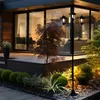 A black metal LED solar candelabra with three lights stands in a garden at dusk, illuminating the surrounding plants and a modern house with large windows. Produktbild-Vorschau