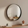 A round wall mirror hangs on a light-colored wall above a wooden shelf with decorative items. Produktbild-Vorschau