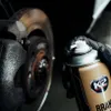 A gloved hand holds a can of brake cleaner next to a car's brake rotor and caliper. Produktbild-Vorschau