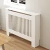 A white radiator cover is shown in a room, placed against a wall and near a window. A wooden table is partially visible in the foreground. Anteprima dell’immagine prodotto