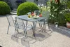 A garden table and chairs set is arranged on a brick patio, surrounded by lush greenery and blooming roses. Produktbild-Vorschau