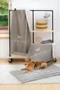A dog is lying on a dog bed in front of a rack with blankets, and a shelf unit with a basket and a stuffed dog. Produktbild-Vorschau