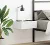 A white floating nightstand is mounted on a white wall next to a bed. A black desk lamp and a white teacup are on top of the nightstand. A large potted plant is to the left of the nightstand. Náhled obrázku produktu
