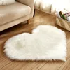 A white heart-shaped shaggy rug is placed on a wooden floor next to a beige sofa. A woven basket filled with books is visible in the background. Anteprima dell’immagine prodotto