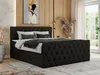 A bedroom scene featuring a black boxspring bed with a tufted headboard and drawers, styled with pillows and a throw blanket. The background includes wood paneling, a piece of art, and a large window with sheer curtains. Produktbild-Vorschau