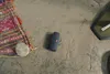 A JBL speaker and a stone are placed on a sandy surface next to a patterned cloth. Produktbild-Vorschau