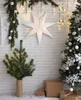 A festive Christmas scene with a white brick wall background, decorated with a large paper star, a small Christmas tree, and a larger decorated Christmas tree. Podgląd zdjęcia produktu