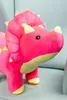 A pink plush dinosaur toy with yellow accents sits on a light gray couch. Voir les images du produit