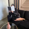 A person wearing a black hooded blanket is reading a book while sitting on a black couch. Náhled obrázku produktu