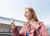 A young woman is on a train platform, smiling and looking at her phone while wearing earbuds. Anteprima dell’immagine prodotto