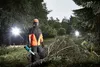 A person in protective gear holding a chainsaw in a forest with bright lights in the background. Náhled obrázku produktu