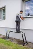 A person is using a multi-purpose ladder with a platform to work on the exterior of a building near a window. Náhľad obrázku produktu