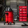 A red industrial vacuum cleaner with a black hose and wand is shown in a warehouse-like setting. Náhľad obrázku produktu