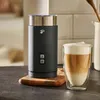 A black electric milk frother next to a glass of latte macchiato on a countertop. Produktbild-Vorschau