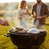 A group of people are having a barbecue in a grassy field during sunset. Burgers are being grilled on a barbecue. Anteprima dell’immagine prodotto