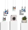A Fujifilm instax SQUARE SQ 1 camera is displayed on a white pedestal, surrounded by hanging photos and a potted plant. Produktbild-Vorschau