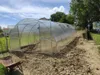 A greenhouse is set up in a garden with a wheelbarrow in the foreground. Produktbild-Vorschau