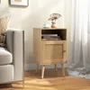 A wooden nightstand with a rattan door is placed next to a sofa in a living room setting. Produktbild-Vorschau