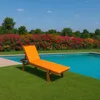A chaise lounge with an orange cushion sits on a grassy lawn next to a swimming pool, with a colorful hedge of flowers in the background. Anteprima dell’immagine prodotto