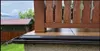 A close-up view of a gutter system and a wooden railing on a balcony or deck, with a small building and trees in the background. Produktbild-Vorschau