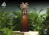 A perfume bottle with a palm tree-like cap, displayed in a lush green jungle setting. Voir les images du produit