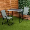 A camping table and two chairs set up on artificial grass in an outdoor setting with a wooden fence and greenery in the background. Produktbild-Vorschau