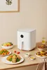 A white air fryer is displayed on a wooden table with various dishes of food around it. Podgląd zdjęcia produktu