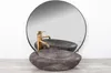 A stone sink with a gold faucet is placed on a wooden surface in front of a large round mirror. Produktbild-Vorschau