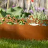 A close-up of a rusty metal garden border with a butterfly design, placed in green grass with blurred flowers and foliage in the background. Produktbild-Vorschau