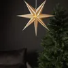A paper star lantern hangs in front of a dark wall, next to a Christmas tree and a dark cushion. Podgląd zdjęcia produktu