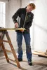 A man is sanding a piece of wood on a sawhorse in a room with patterned wallpaper and wooden floors. Produktbild-Vorschau