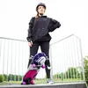 A young woman in a black hoodie and leggings stands with a skateboard at a skate park. Voir les images du produit