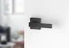 A black door handle is mounted on a white wall. The background on the left is blurred and shows a room interior. Produktbild-Vorschau