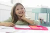 A young girl is sitting at a table with art supplies, looking up and smiling. Anteprima dell’immagine prodotto