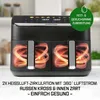 A black dual-basket air fryer with glowing heating elements inside the baskets, set against a blurred kitchen background. Produktbild-Vorschau