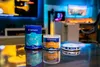 Three spindles of optical discs (CD-R, DVD-R, BD-R) are displayed on a reflective surface in front of a blurred background of a room with screens and lighting. Produktbild-Vorschau