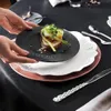 A person's hands are placing a small black plate with food on top of a stack of larger plates on a black tablecloth. Produktbild-Vorschau