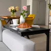 A black C-shaped side table is placed on the armrest of a sofa, holding a coffee cup, a croissant on a plate, and a vase with flowers. Produktbild-Vorschau