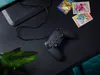 A black gaming controller is placed on a black surface, with some collectible cards and a plant in the background. Náhled obrázku produktu