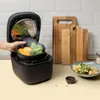 A hand is shown placing a steaming bowl of vegetables into an open rice cooker. In the background, there are wooden cutting boards and a bowl of food. Produktbild-Vorschau