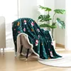 A dark green blanket with a Christmas-themed pattern is draped over a chair in a room with a large plant. Produktbild-Vorschau