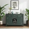 A green sideboard with decorative elements, flanked by potted plants, with artwork and a lamp on top. Produktbild-Vorschau