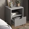 A grey bedside table with an open drawer, displaying books and a folded blanket. A gold clock and a potted plant are on top. Voir les images du produit