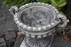 A stone amphora planter sits on a stone base outdoors, surrounded by rocks and foliage. Produktbild-Vorschau