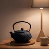 A dark blue cast iron teapot sits on a wooden table next to a lit lamp and a candle. Podgląd zdjęcia produktu