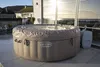 An inflatable hot tub covered with a lid sits on a tiled patio next to a building with large windows overlooking the ocean. Produktbild-Vorschau