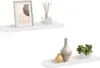 Two white wall shelves are displayed against a white background. The top shelf holds a framed picture and a diffuser bottle with reeds. The bottom shelf displays two vases and a small potted plant. Náhľad obrázku produktu