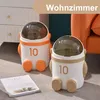 Two astronaut-shaped trash cans are placed on a carpeted floor next to a piece of furniture and an orange chair. A magazine is placed in front of the trash cans. Produktbild-Vorschau